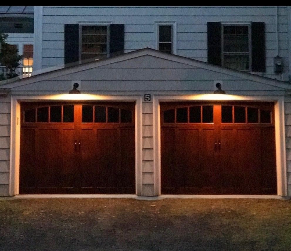Two dark brown garage doors with overhead lights, located in front of a white house with black shutters.