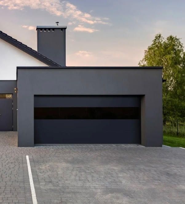 Modern gray garage with a horizontal black stripe, connected to a white house with a chimney, on a brick paved driveway.