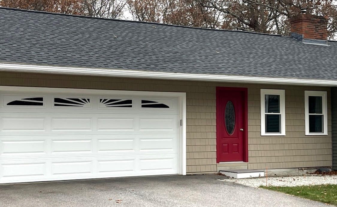 White garage door, red front door, two white windows on a light brown house with dark gray roof.