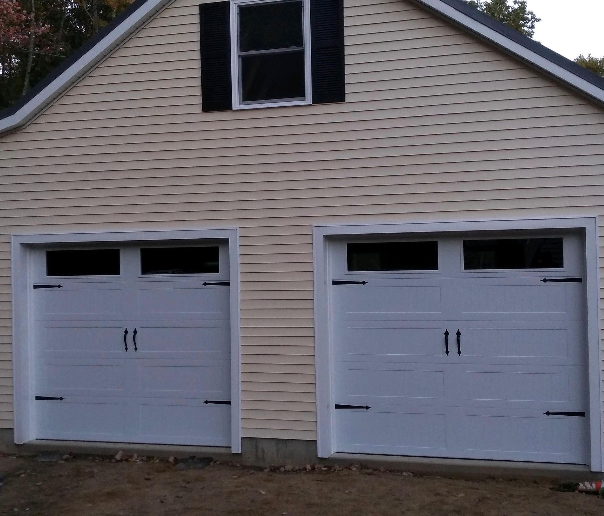 Two white garage doors with black hardware, tan siding, and a small window above.