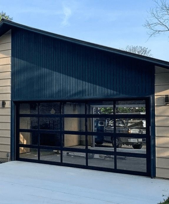 Garage with black-framed glass door, dark blue siding above, light-colored siding on the side, concrete driveway.