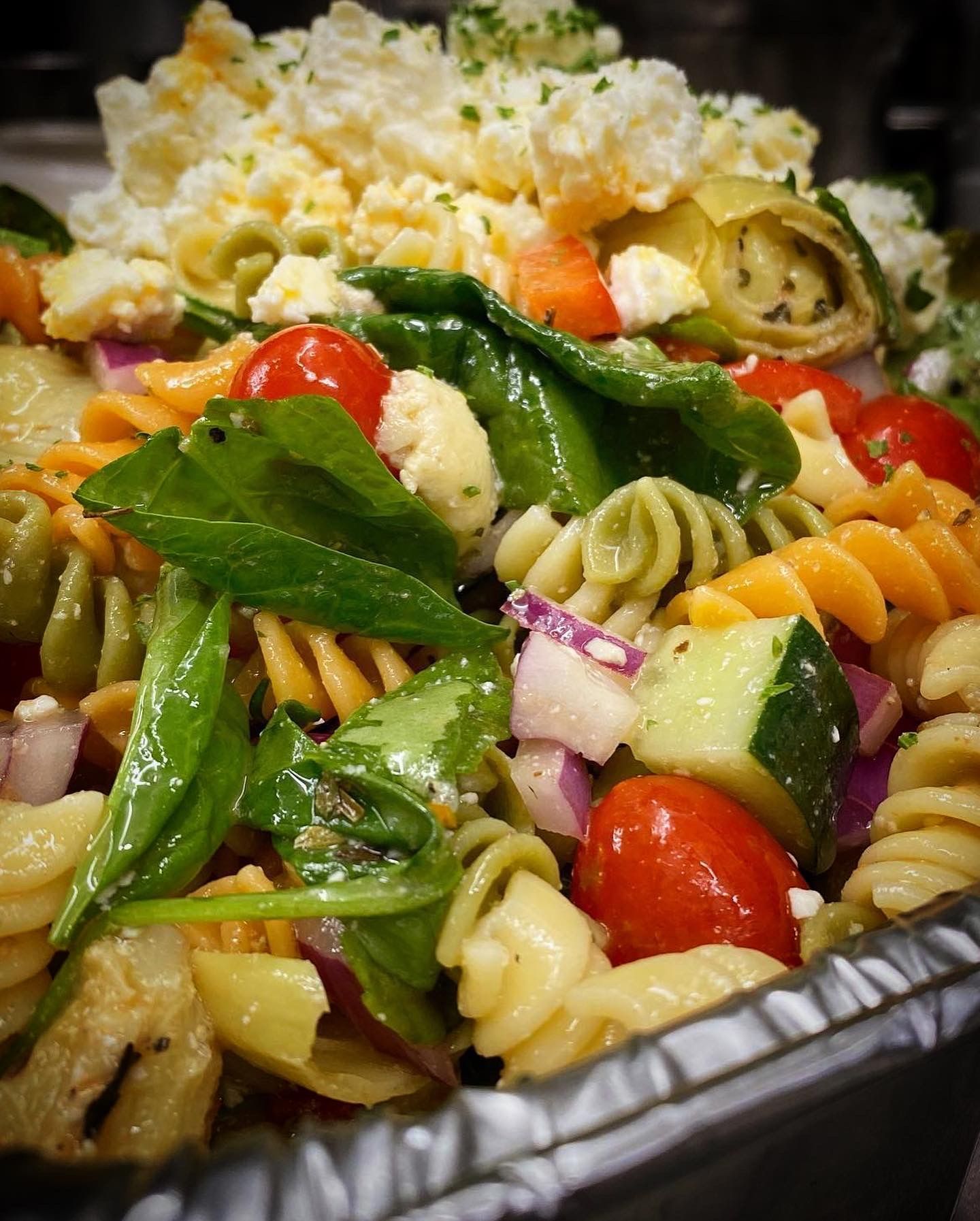 A pasta salad with tomatoes , cucumbers , onions and spinach in a foil container.