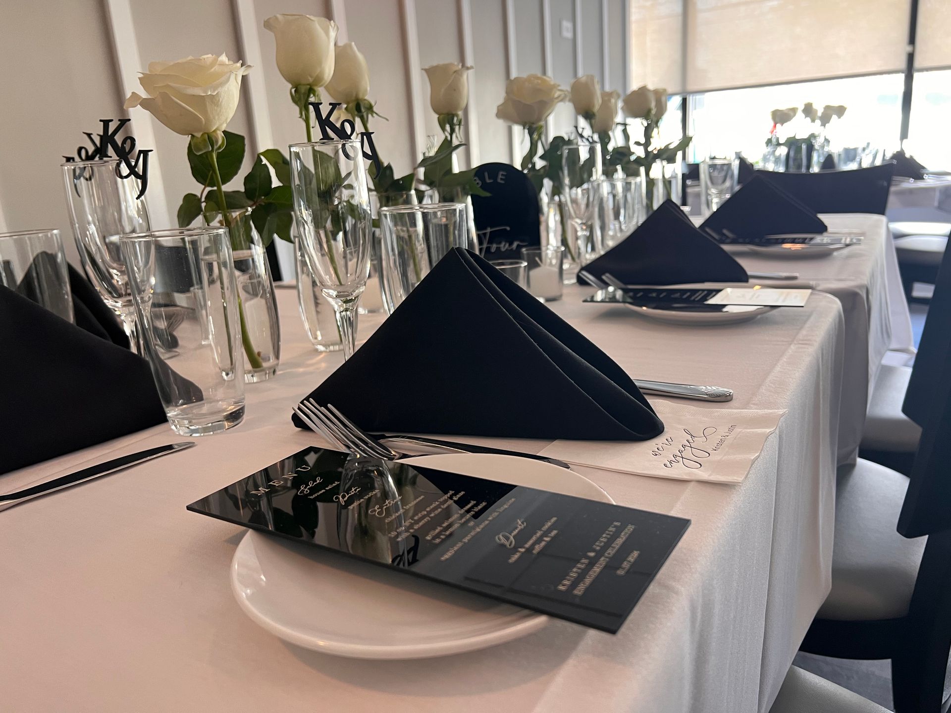 A long table with black napkins , plates , glasses and flowers on it.