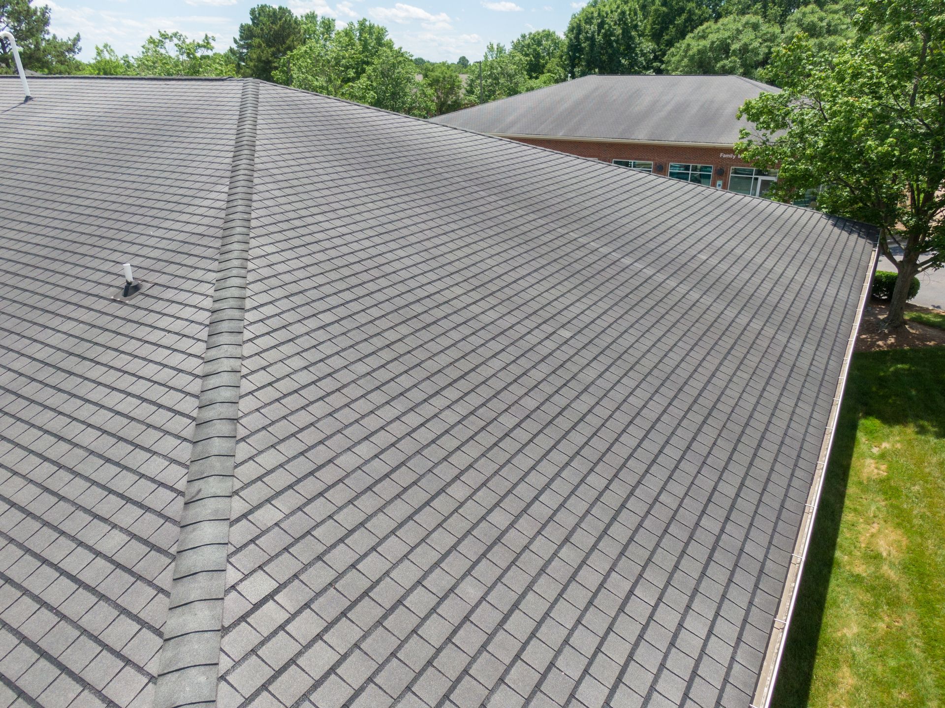 An angled, high-view shot of a gray, textured shingled roof with a central ridge, framed by green trees and lawn.