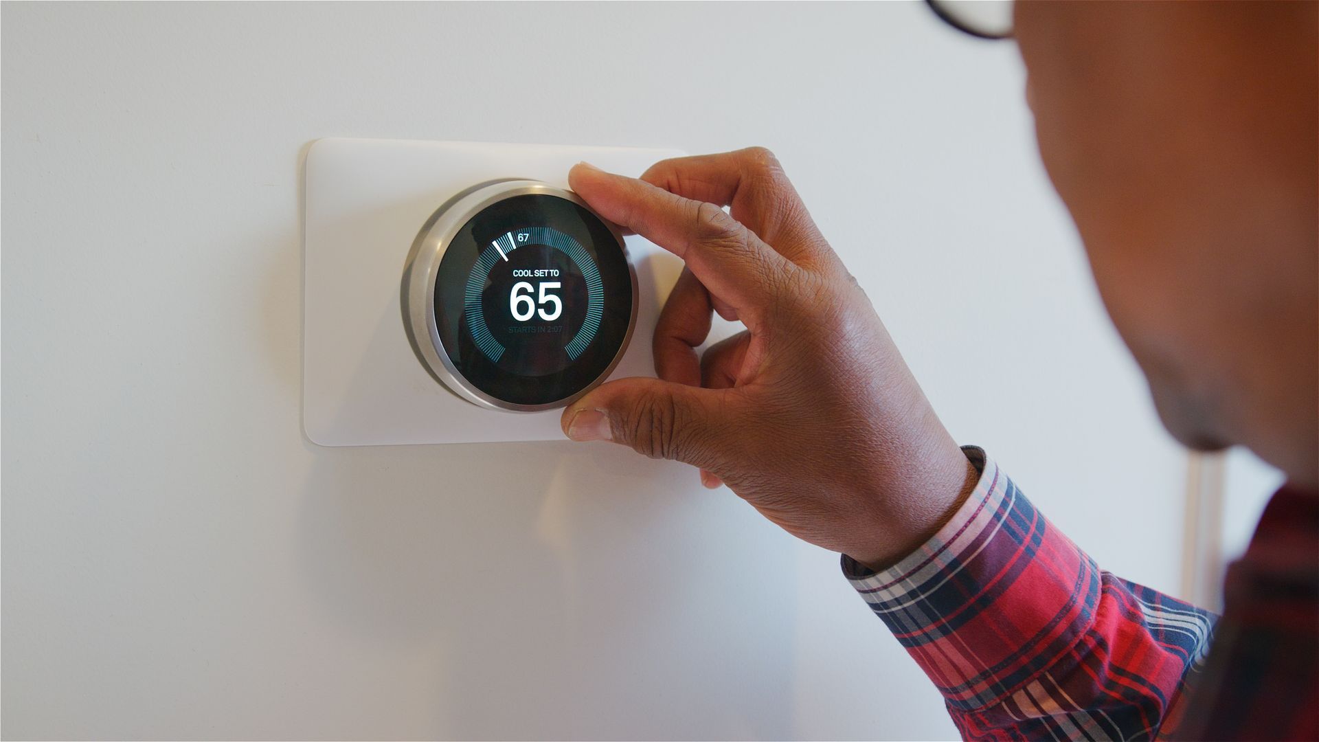 A person in a plaid shirt adjusts the temperature on a circular smart thermostat set to 65 degrees.