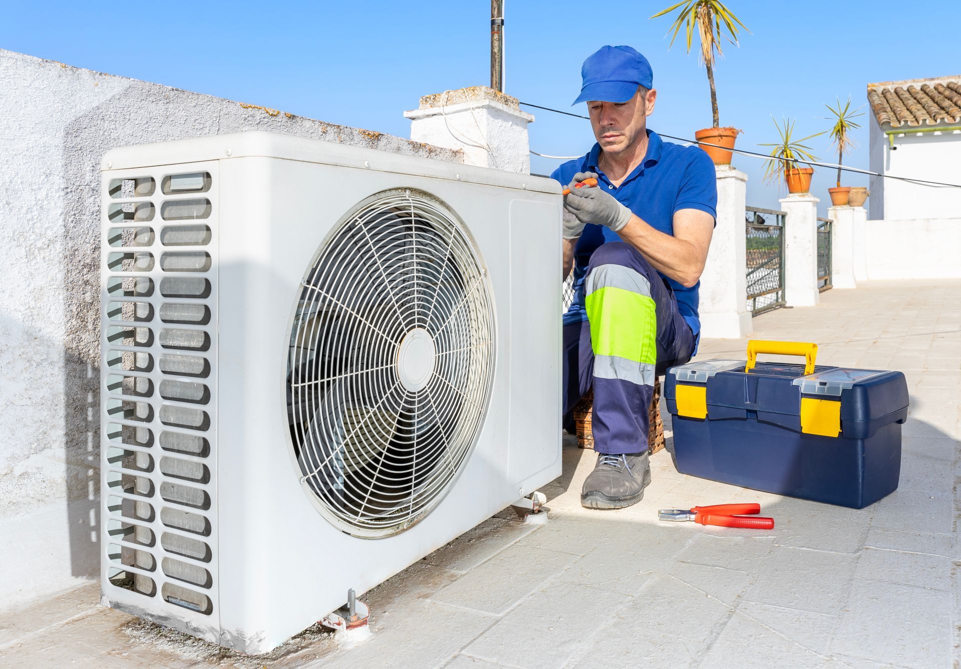 A technician in a blue uniform and cap repairs an outdoor air conditioning unit on a sunny rooftop terrace.
