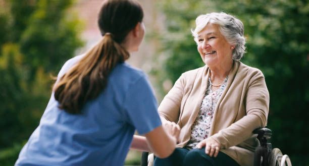 A caregiver holding hands with a senior woman in a wheelchair while smiling.