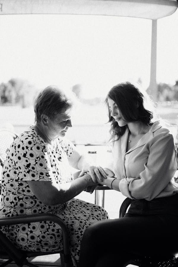 A senior woman in a wheelchair and her caregiver smiling outside in a park.