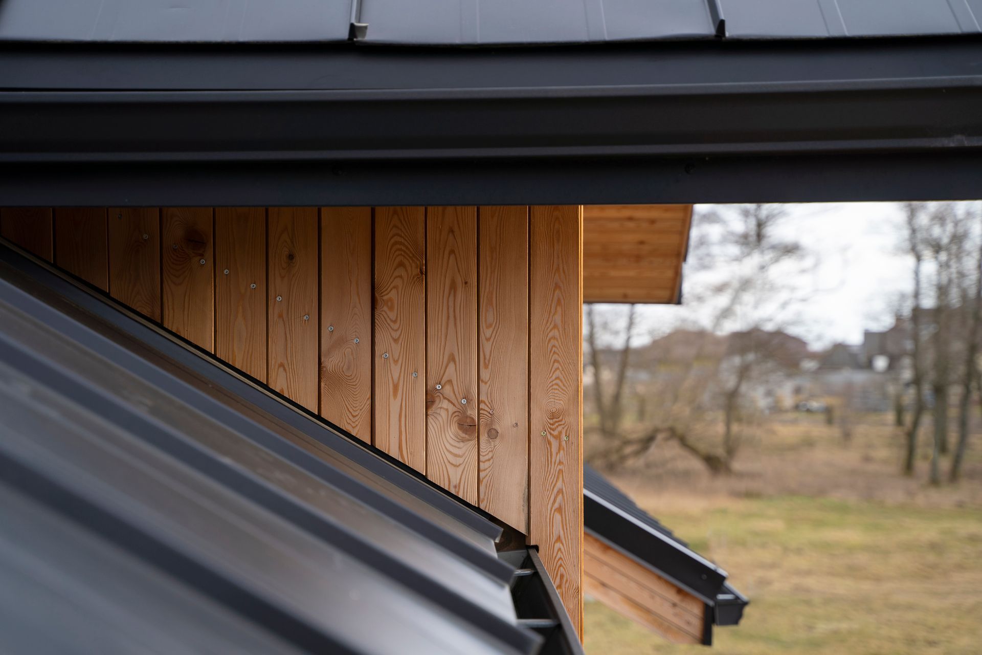 Close-up of wooden siding and black metal roof with a view of trees and houses in the background.