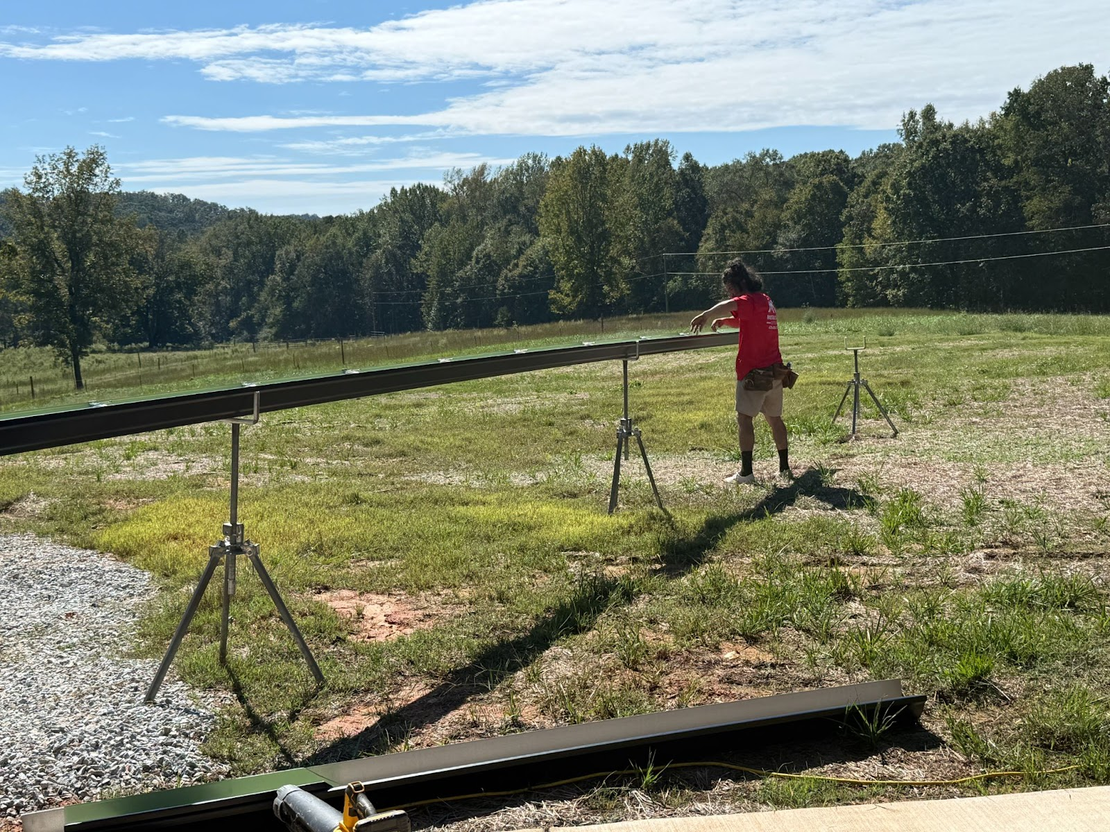 Person in red shirt adjusting equipment in a grassy field with trees and a blue sky in the background.