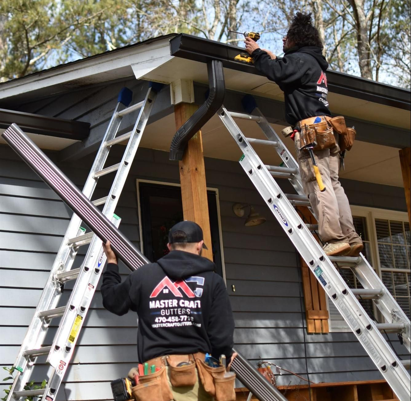 Two workers on ladders repairing a house roof and gutter under an eave