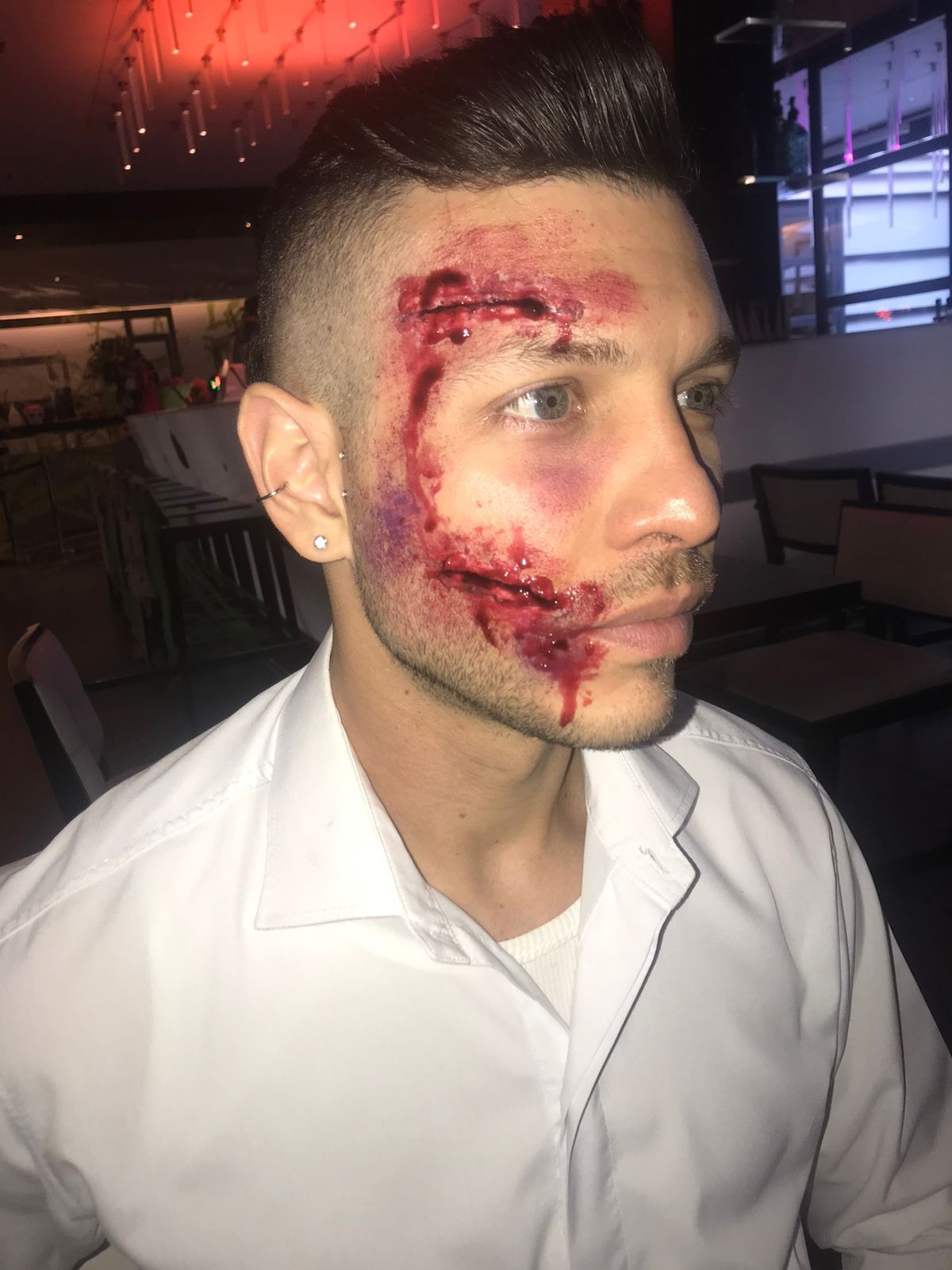 Hombre con laceraciones falsas y hematomas en la cara, vestido con camisa blanca.