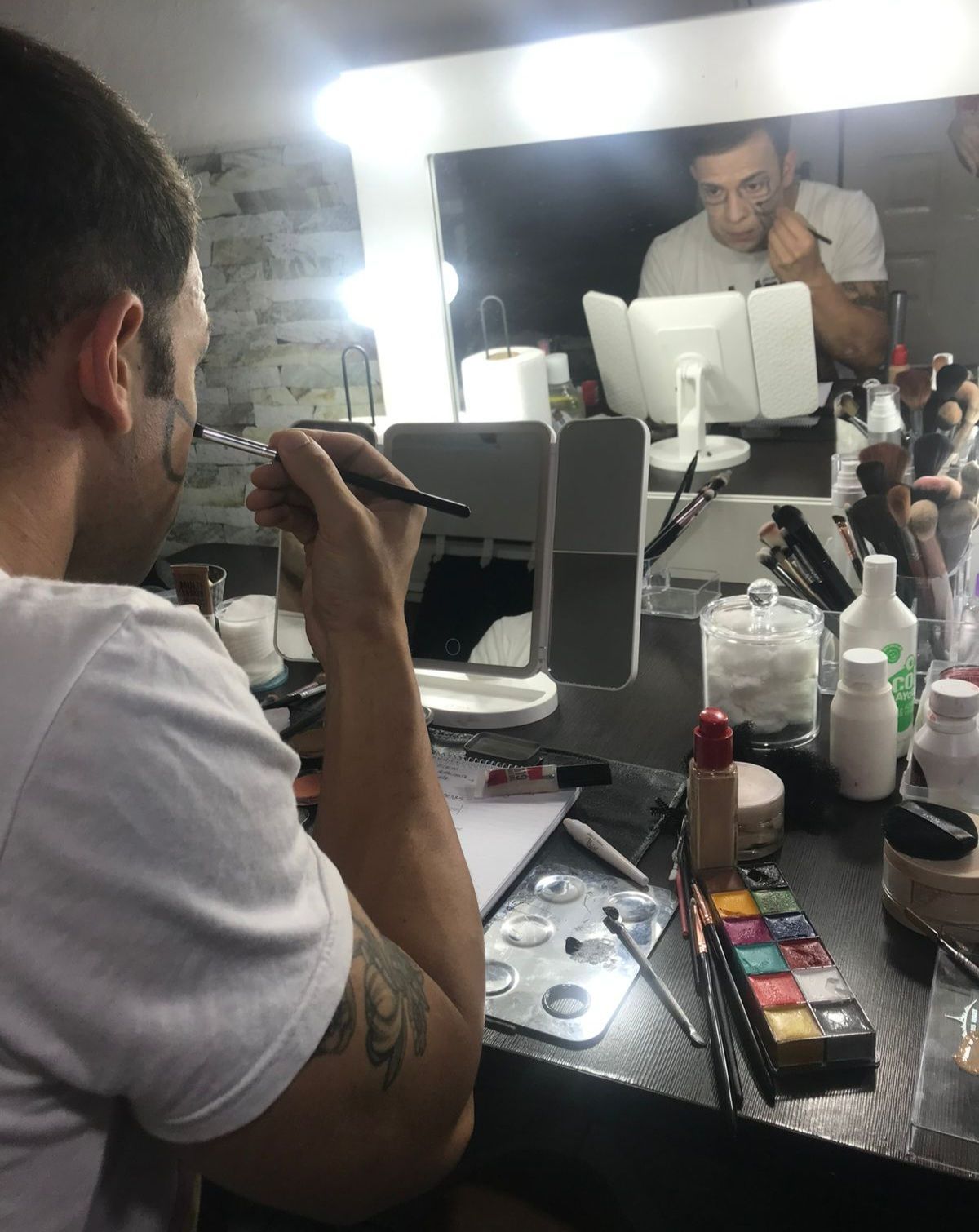 Hombre maquillándose frente a un espejo de tocador, con reflejo visible.