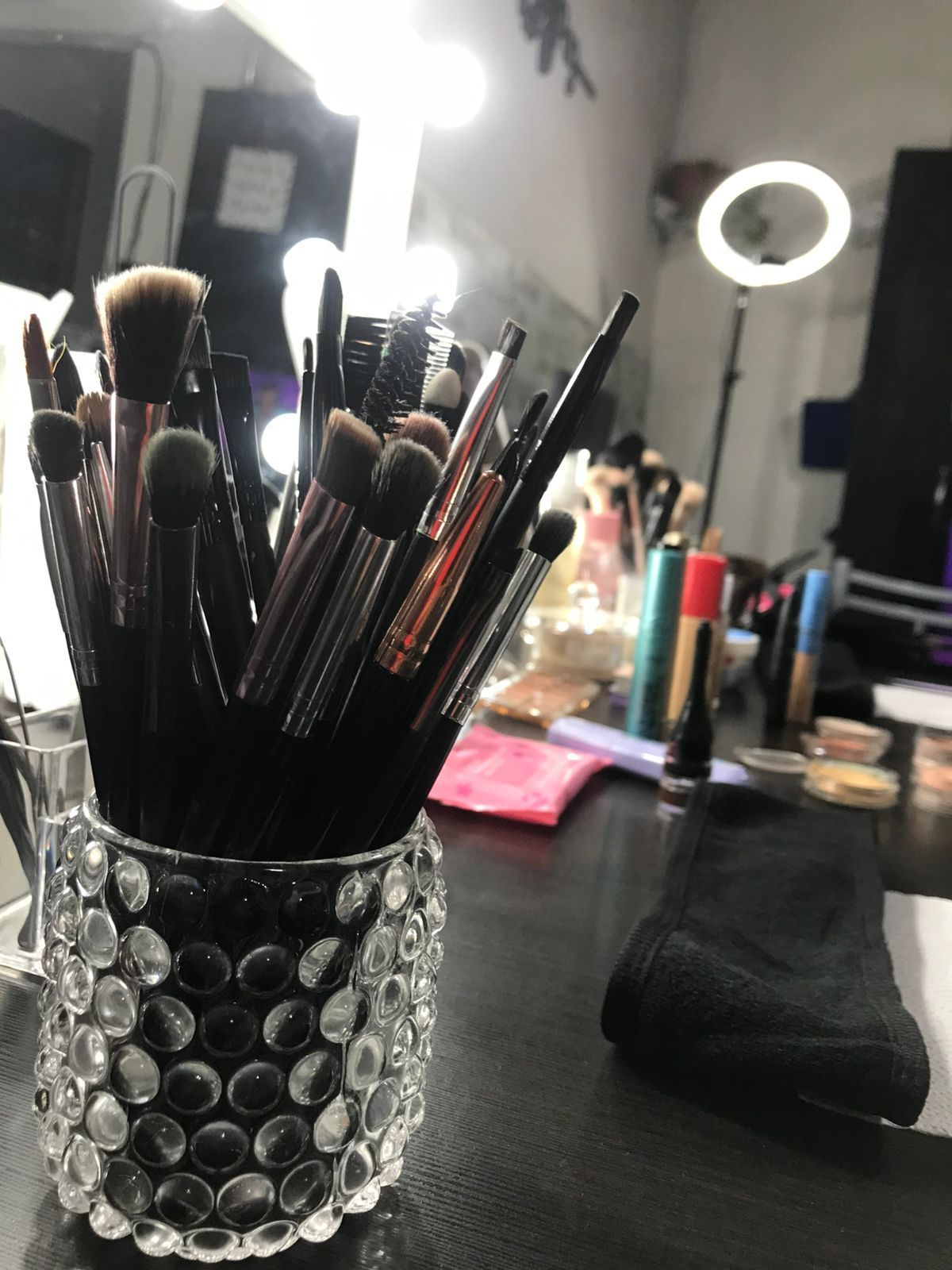 Pinceles de maquillaje en un soporte de cristal sobre una mesa, con un espejo iluminado y un anillo de luz en el fondo.