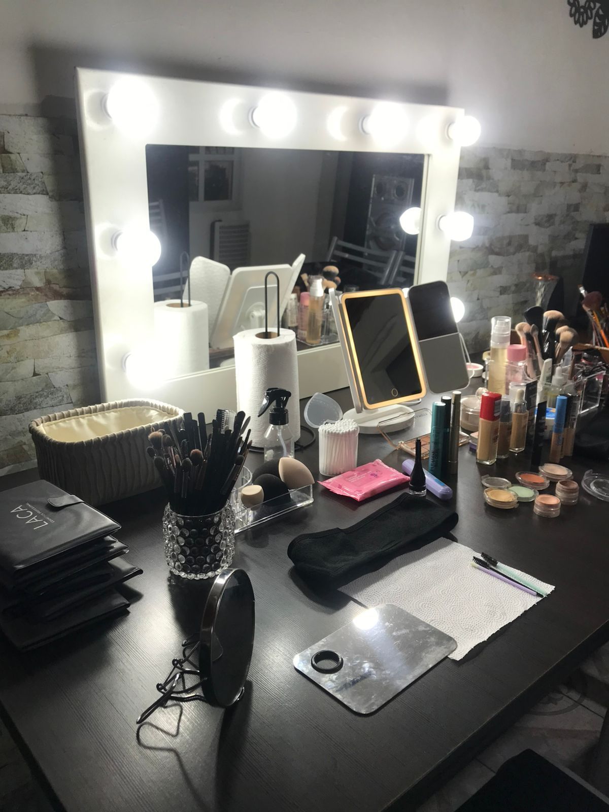 Tocador de maquillaje con espejo iluminado, pinceles y diversos productos cosméticos sobre una mesa oscura.