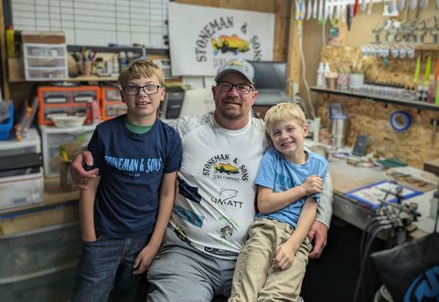 Man with two young sons smiling in a workshop; sign: Stoneman & Sons, fishing lures in background.