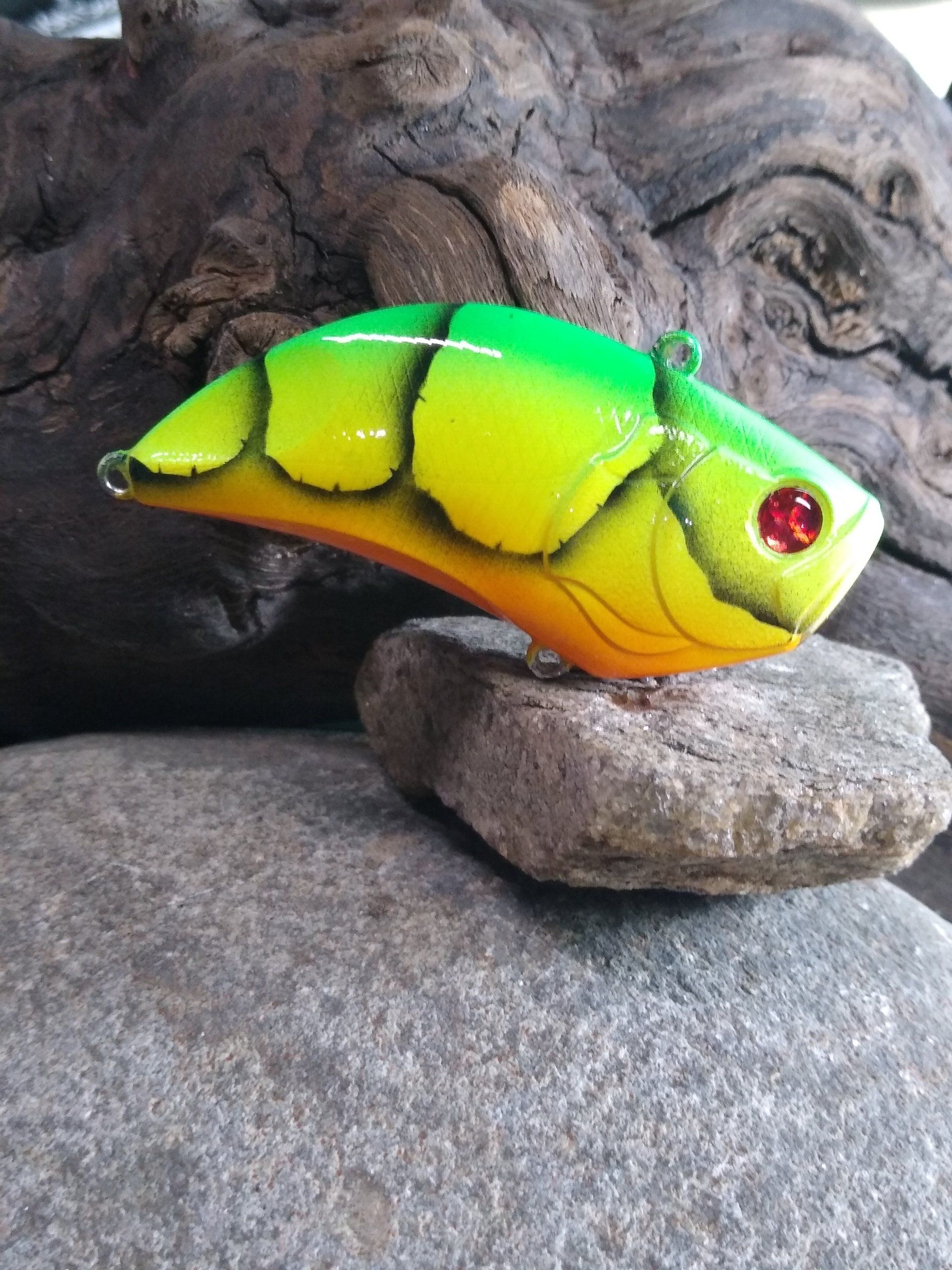 Green and yellow fishing lure with red eyes, resting on a gray rock, against a dark wooden background.