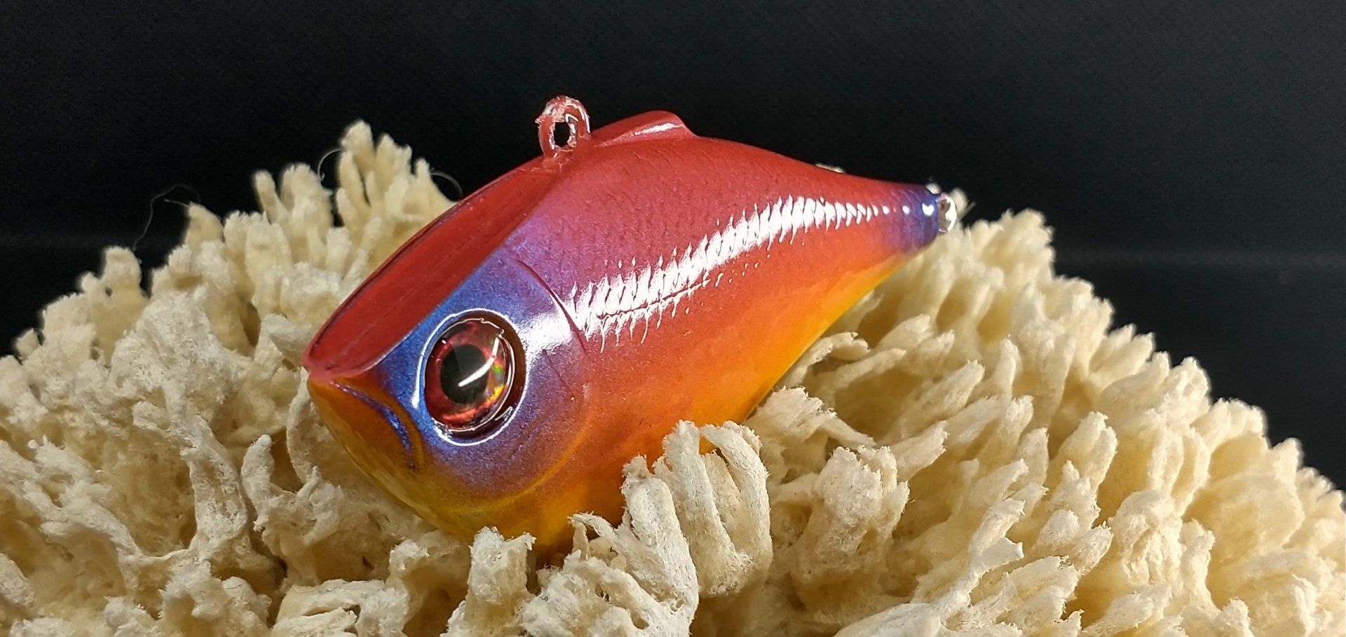 A colorful fishing lure, red, orange, and purple, resting on a textured, beige surface.