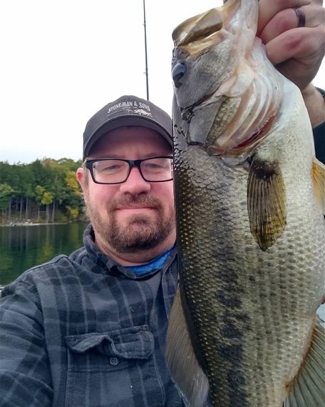 Man holding a large bass fish he caught near a body of water, smiling, wearing glasses and a hat.