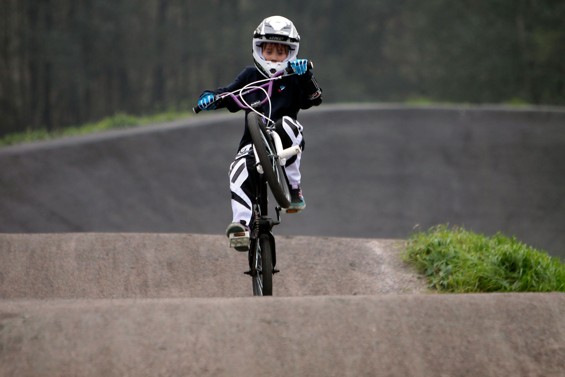 A person wearing a helmet and protective gear rides a BMX bike on a dirt track, balancing on the rear wheel in a wheelie.