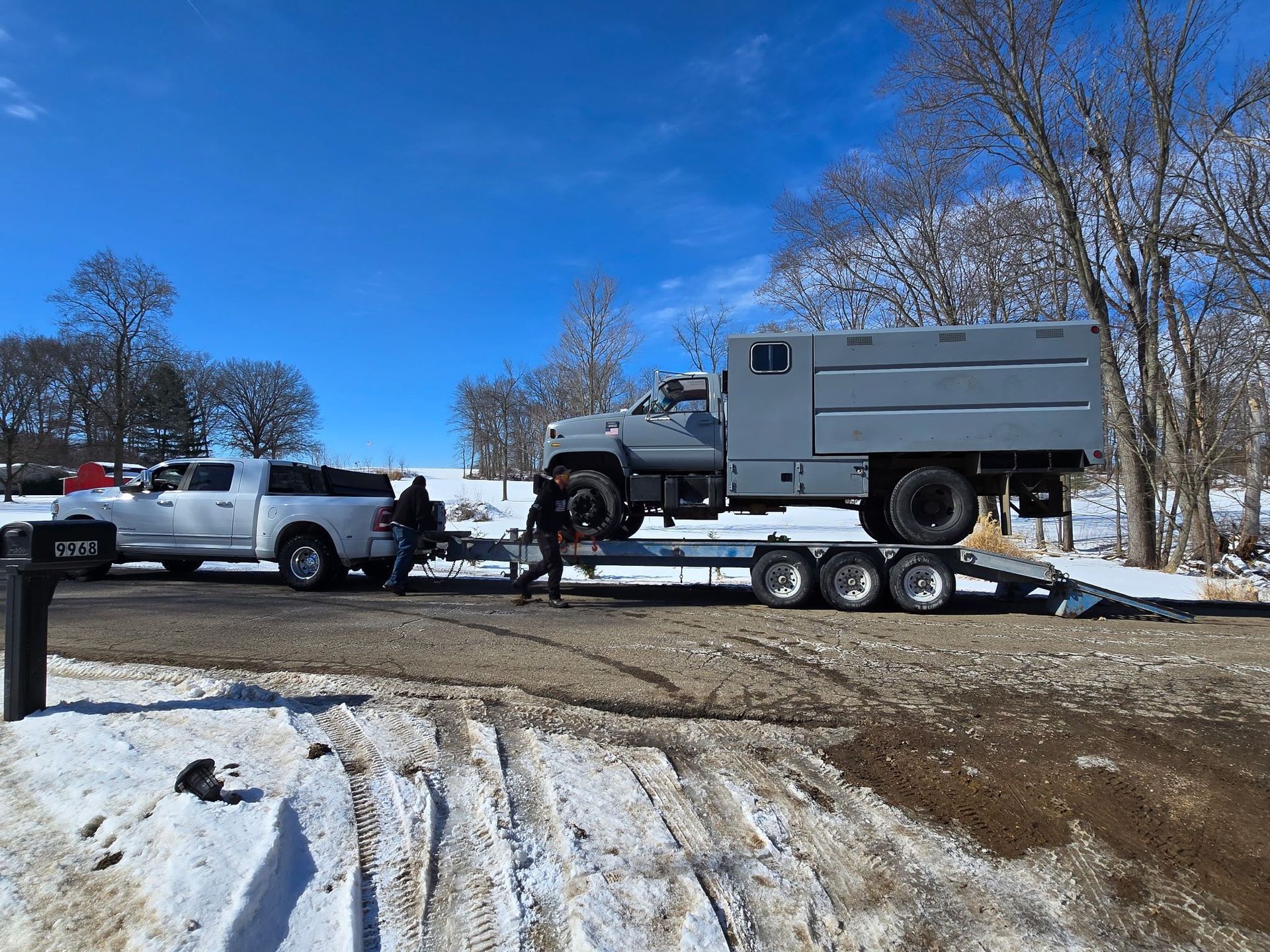 A white pickup truck towing a flatbed trailer with a large gray service truck on it in a snowy, outdoor setting.