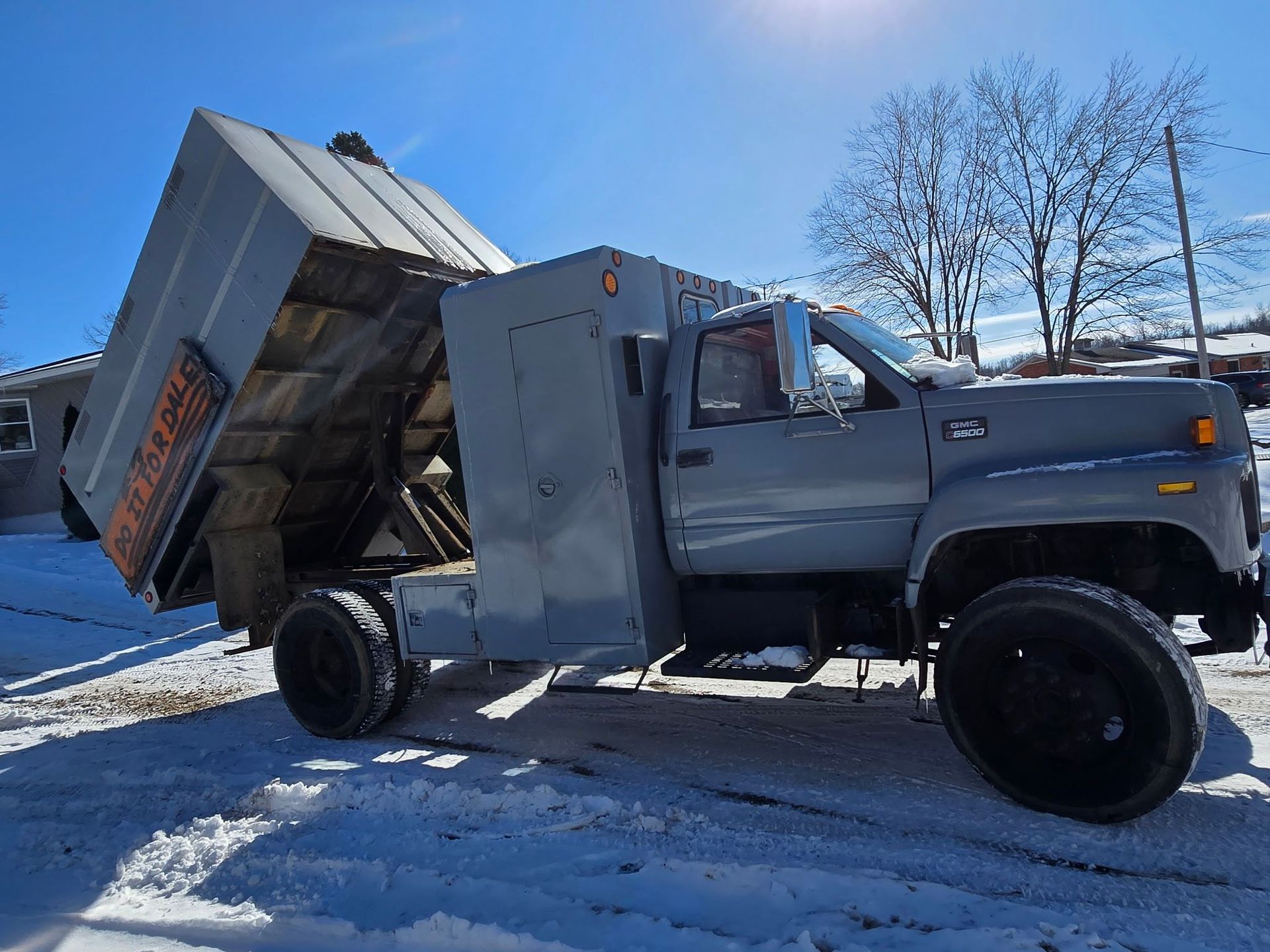 A grey dump truck with a raised bed parked on a snow-covered surface under a bright blue sky.