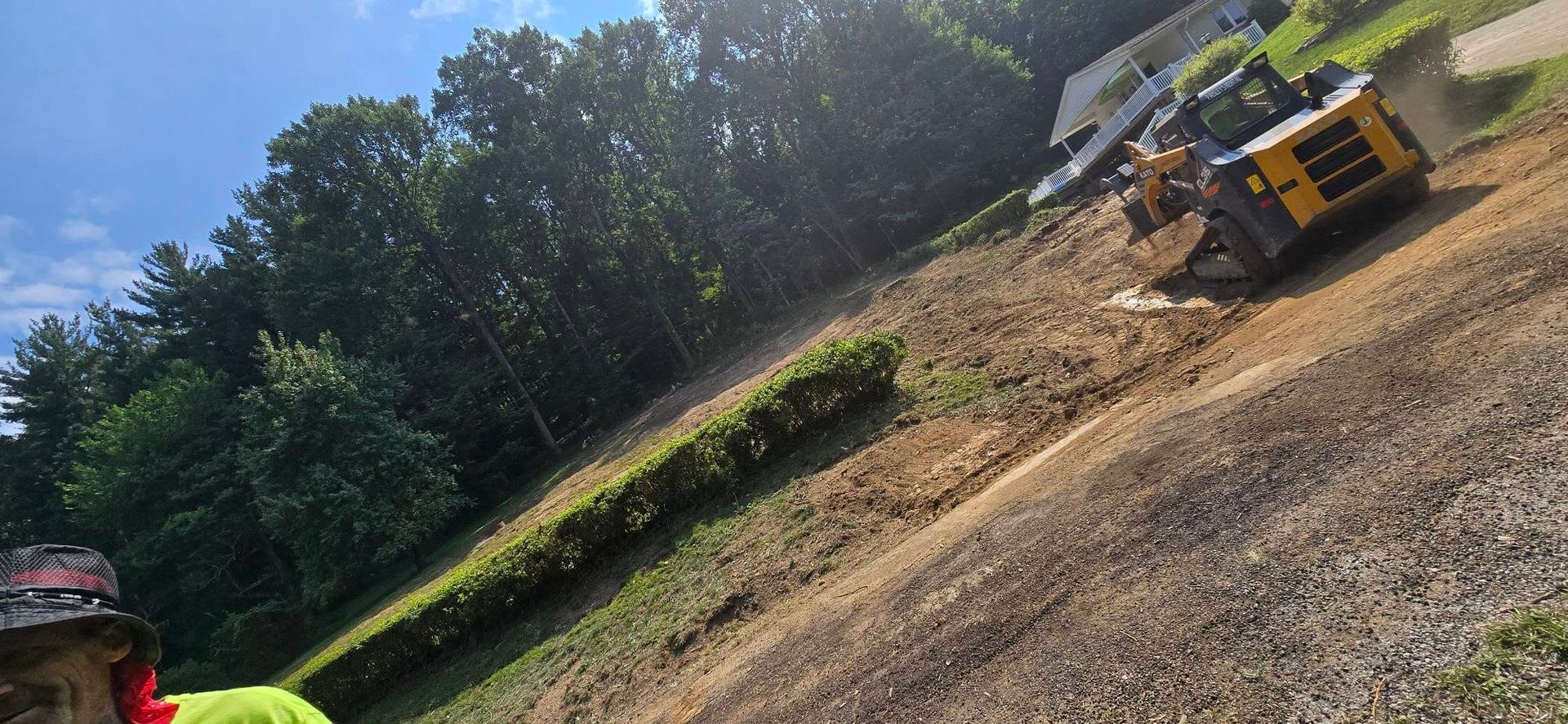 A yellow construction loader sits on a dirt lot cleared of trees near a forest edge, with a person in view.