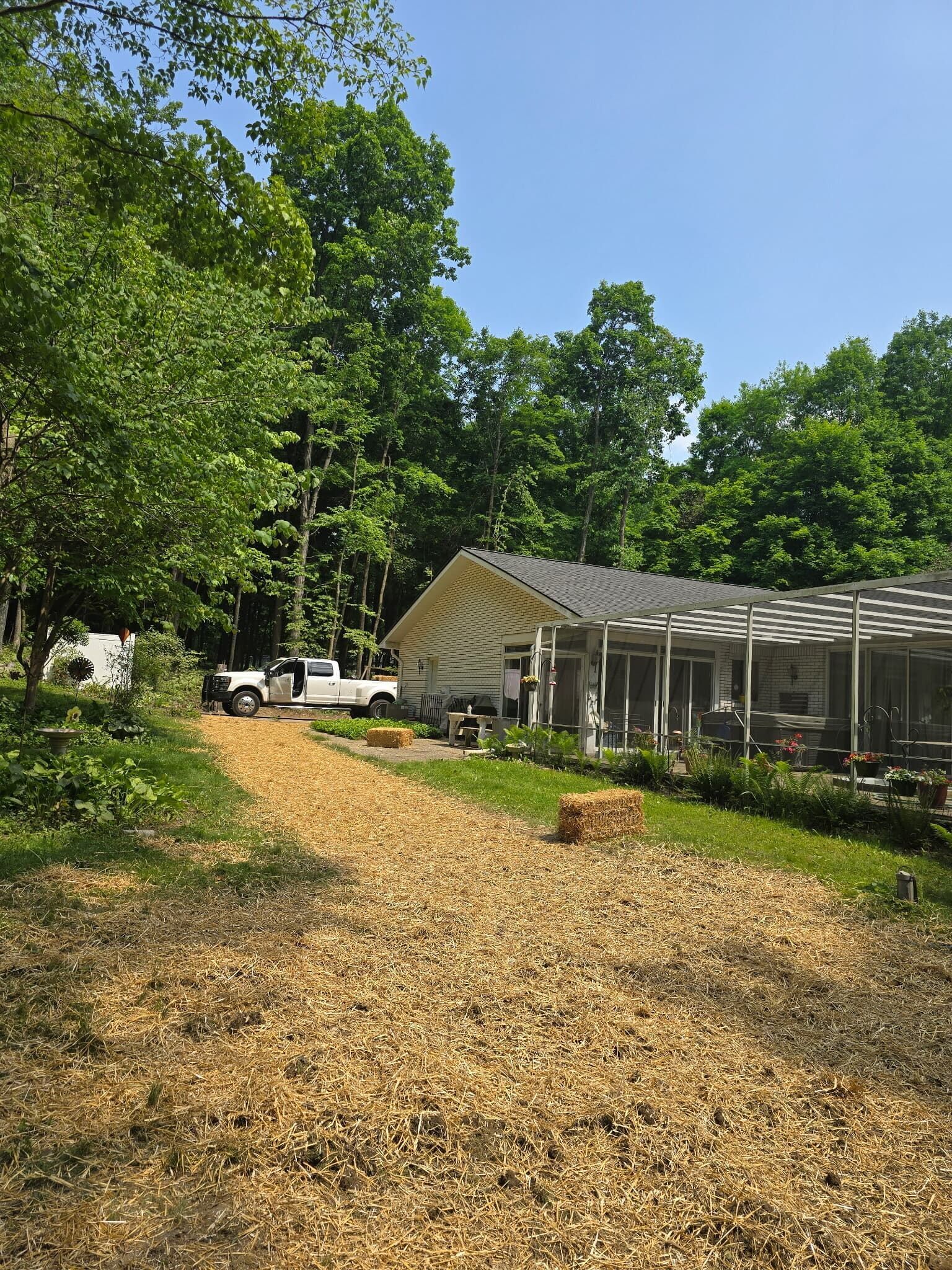 A wood-chip path leads toward a white house with a screened porch and a truck parked beside a wooded area.