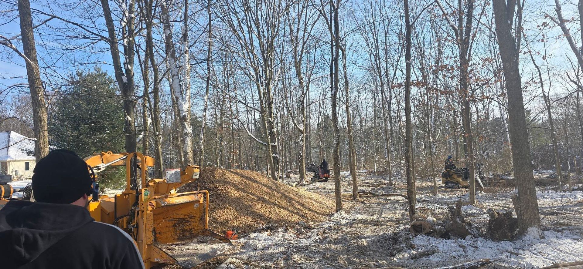 A person in a black jacket stands by yellow construction equipment near a pile of dirt in a snowy, wooded area.