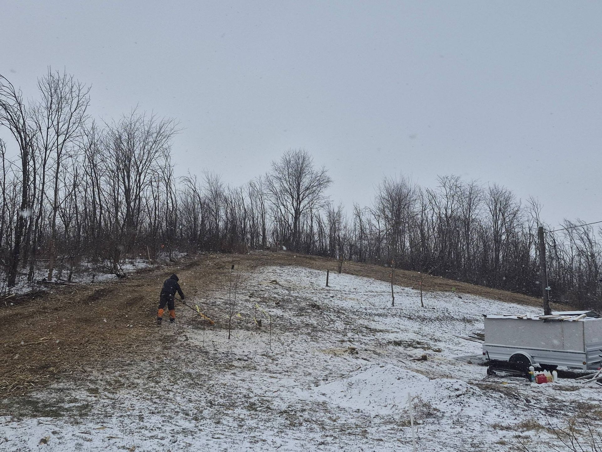 A person works on a snowy, grassy hillside near a parked trailer under a gray sky.