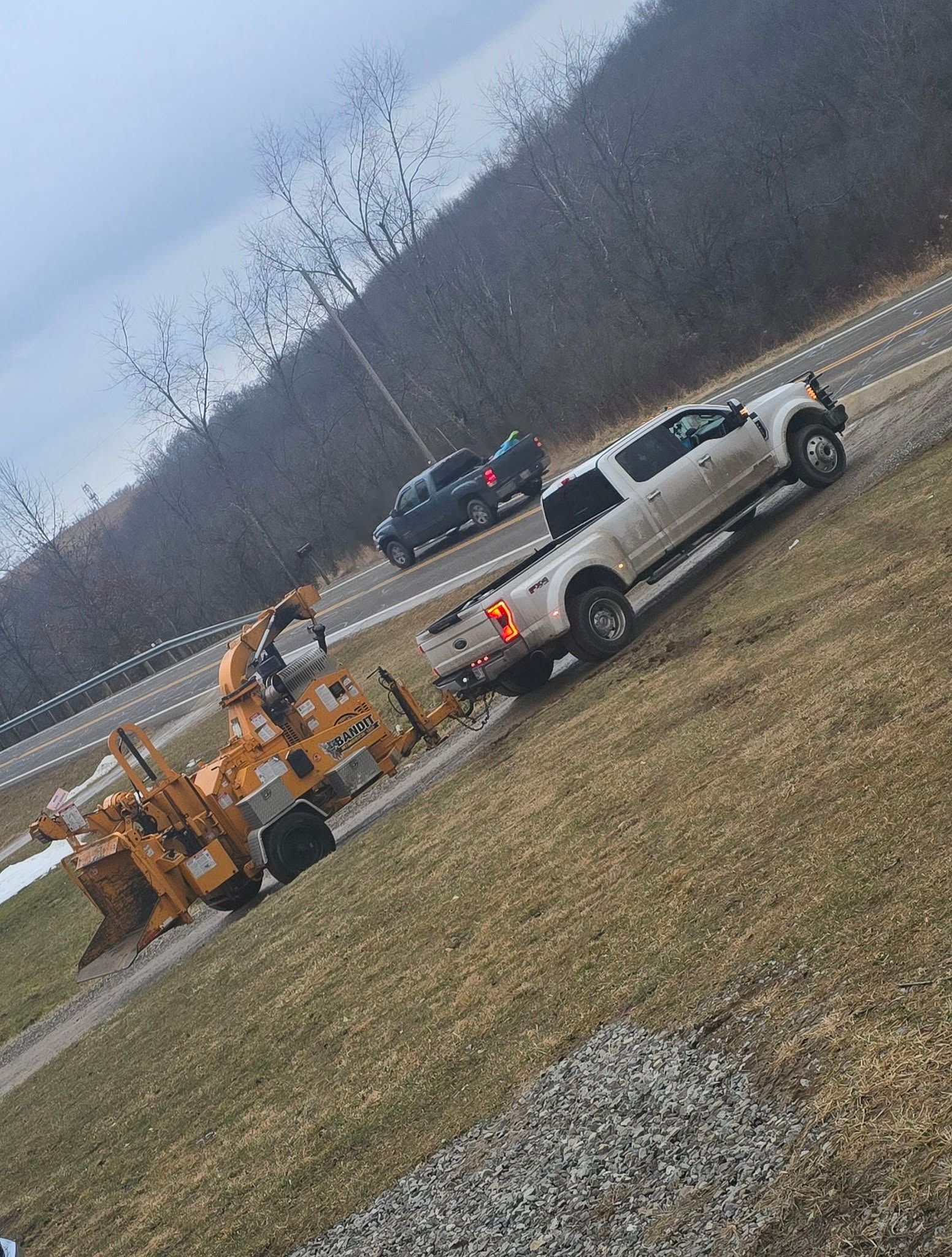 A white pickup truck parked on grass tows a large yellow wood chipper, with a dark SUV driving on a road behind it.
