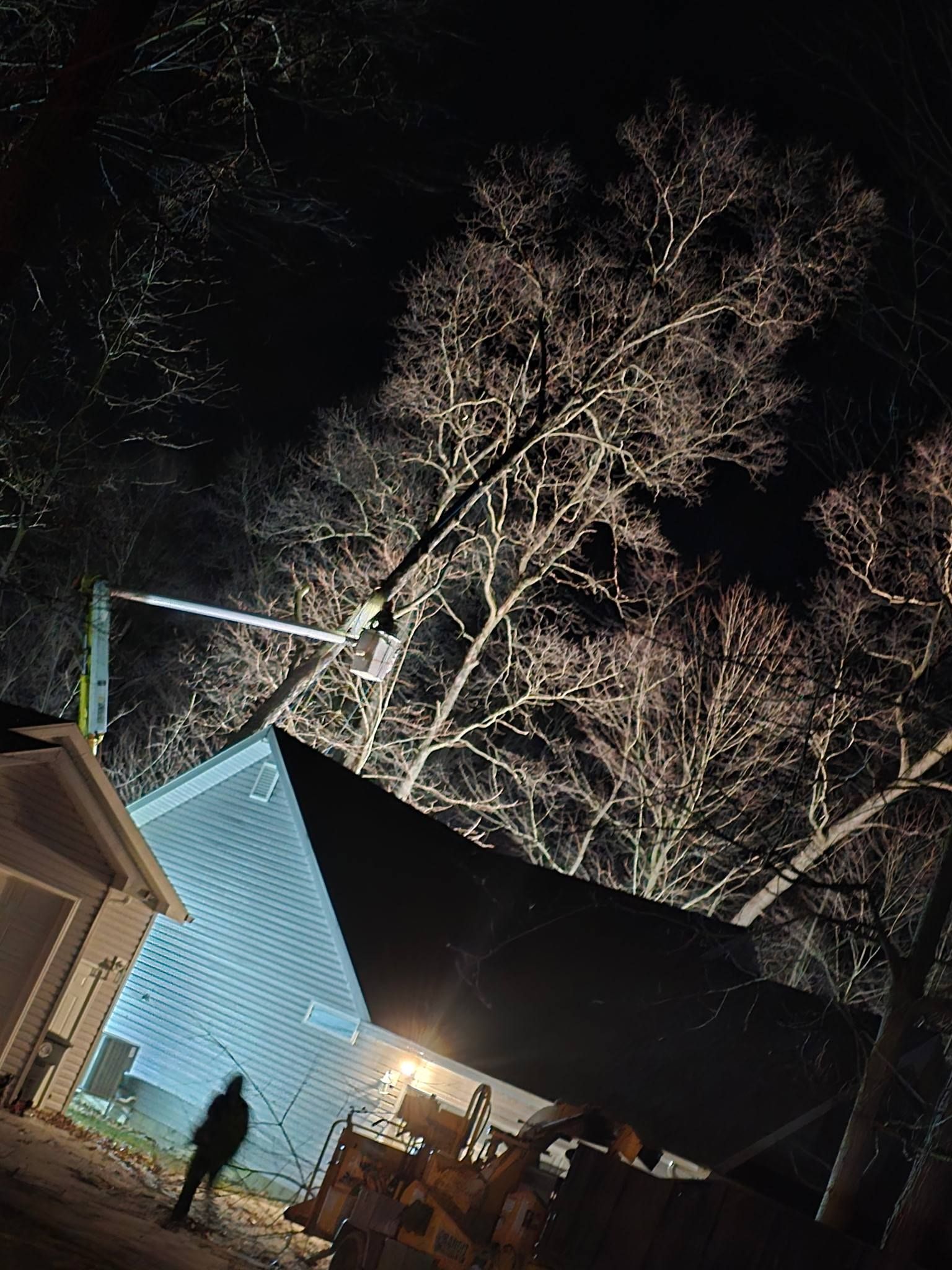 At night, a crane lifts a house roof section into place near trees, illuminated by bright construction lights.