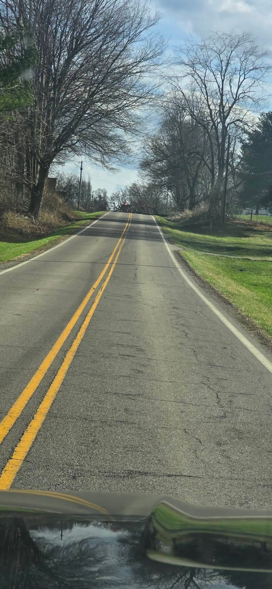 A two-lane road with yellow center lines leads up a gentle hill through a landscape of bare trees under a cloudy sky.