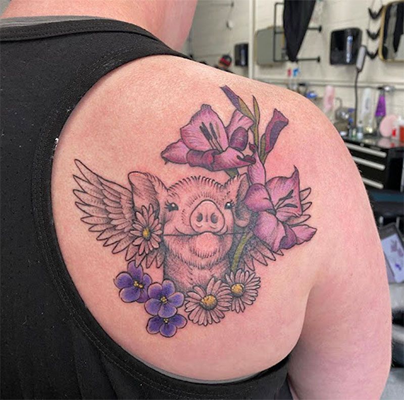 Tattoo of a pig with wings and flowers on a person's shoulder. Pink, purple, and white colors.