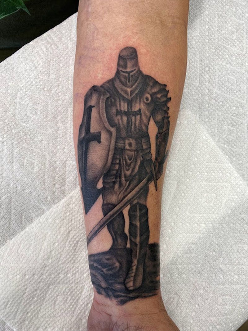 Tattoo of a knight in full armor standing on a rocky surface, holding a shield and sword.