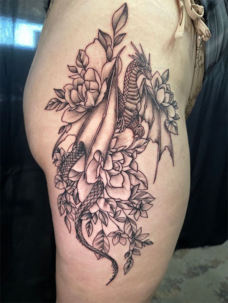 A black and white tattoo of a dragon surrounded by flowers on a woman 's thigh.