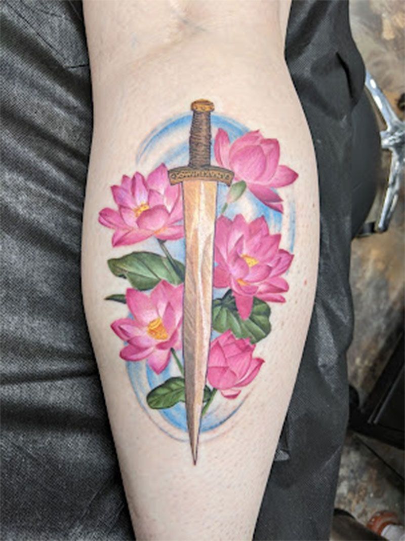 Tattoo of a bronze sword through pink lotus flowers, surrounded by blue strokes on a leg.