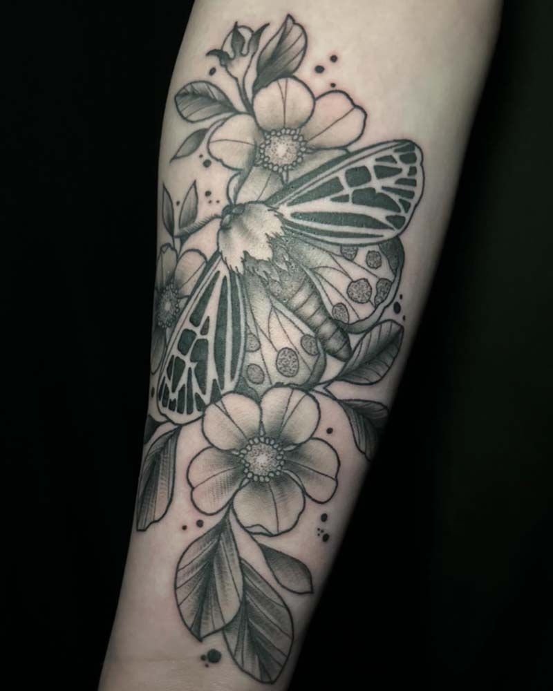 A black and white tattoo of a moth and flowers on a person 's arm.