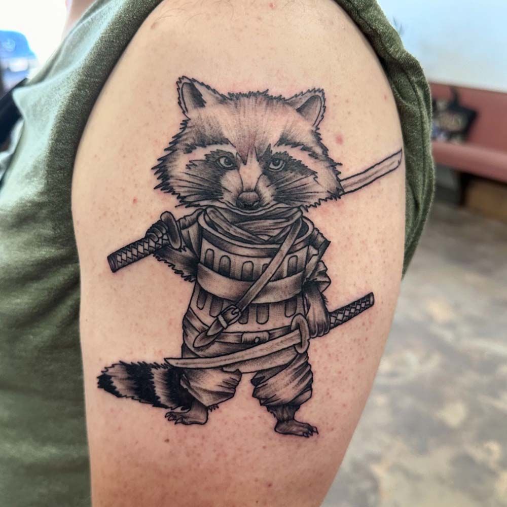 Tattoo of a raccoon dressed as a samurai, holding two katanas, on an arm.