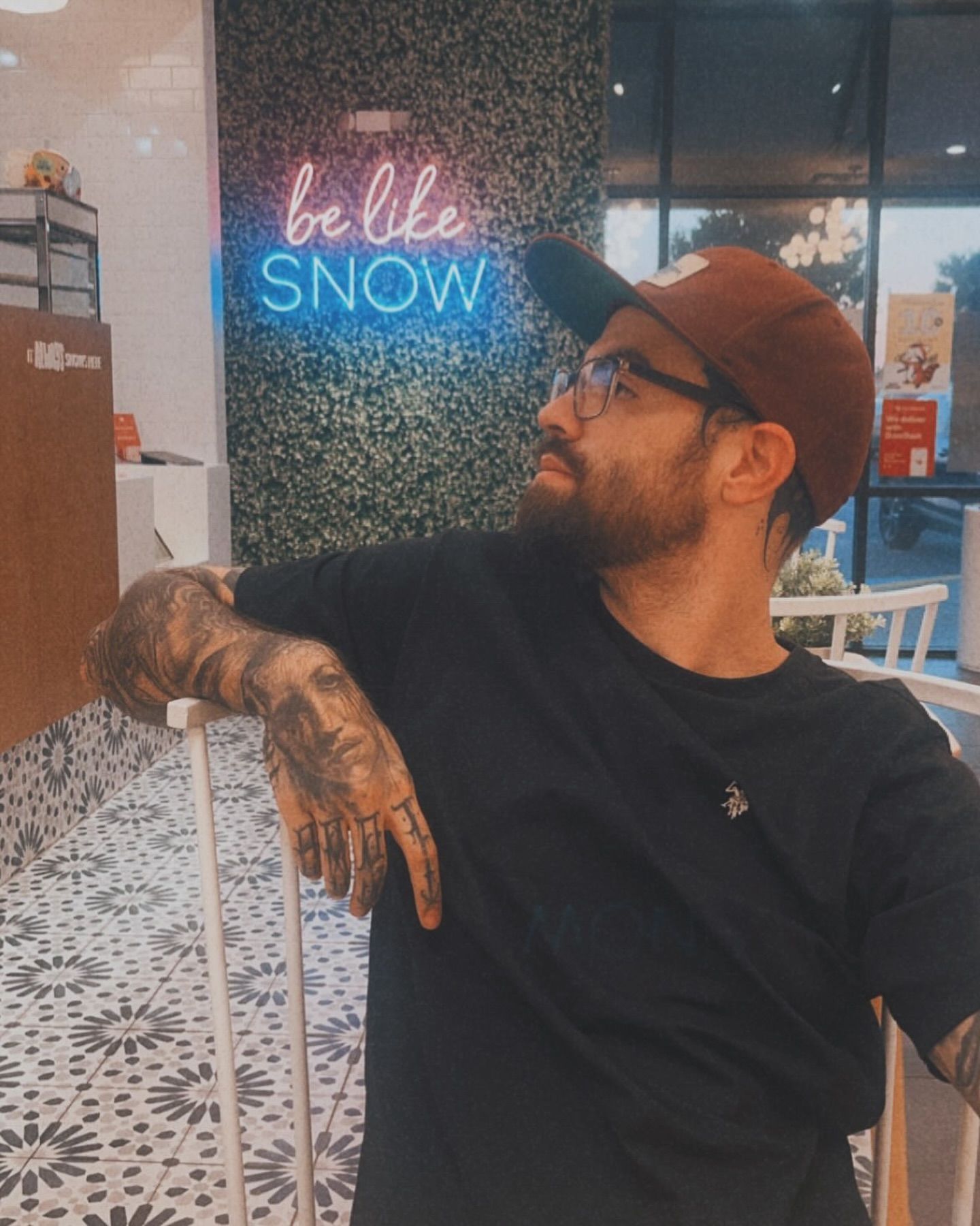 A man leaning on a railing in front of a neon sign that says be like snow