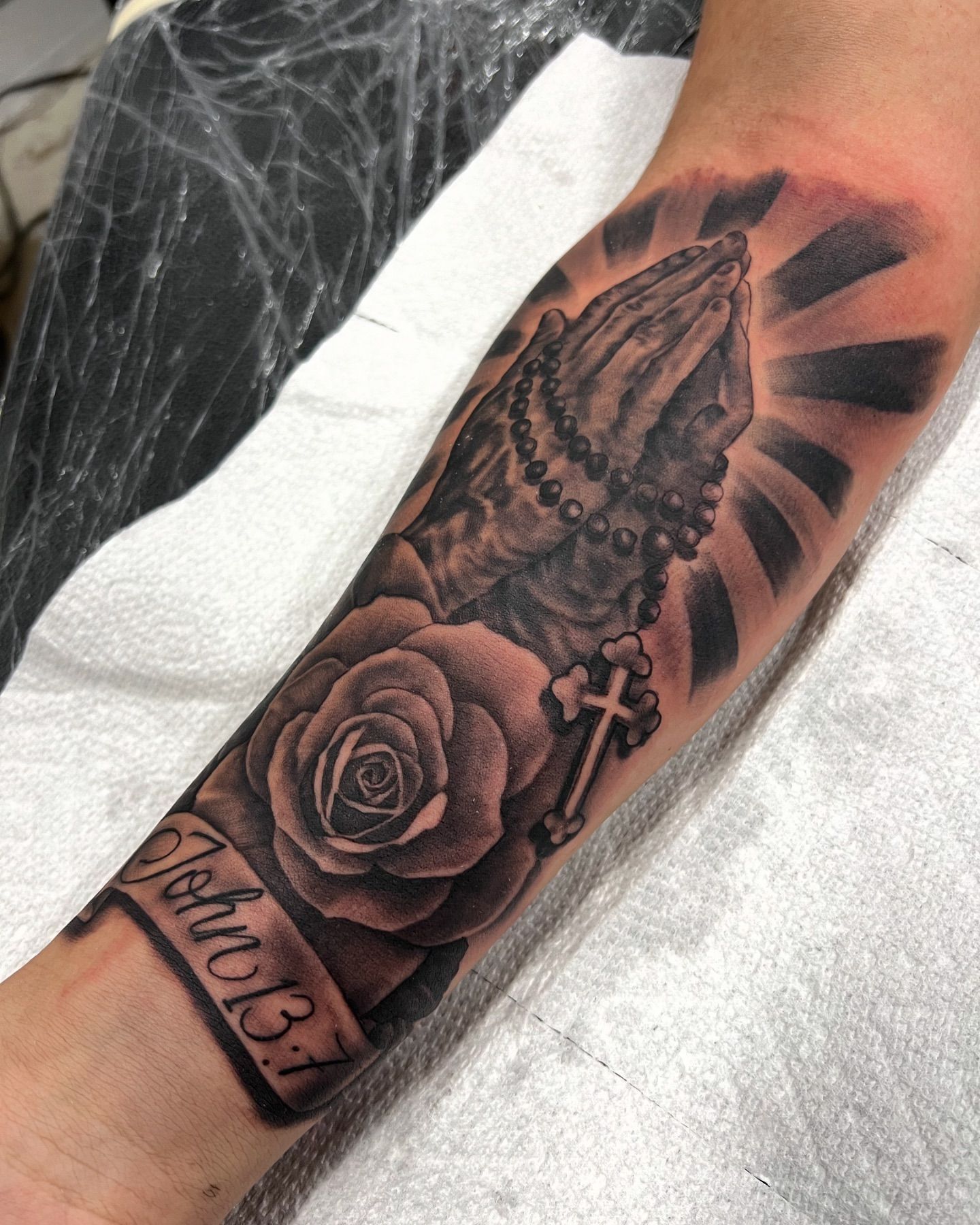 A man has a tattoo of a praying hand with roses and a cross on his forearm.