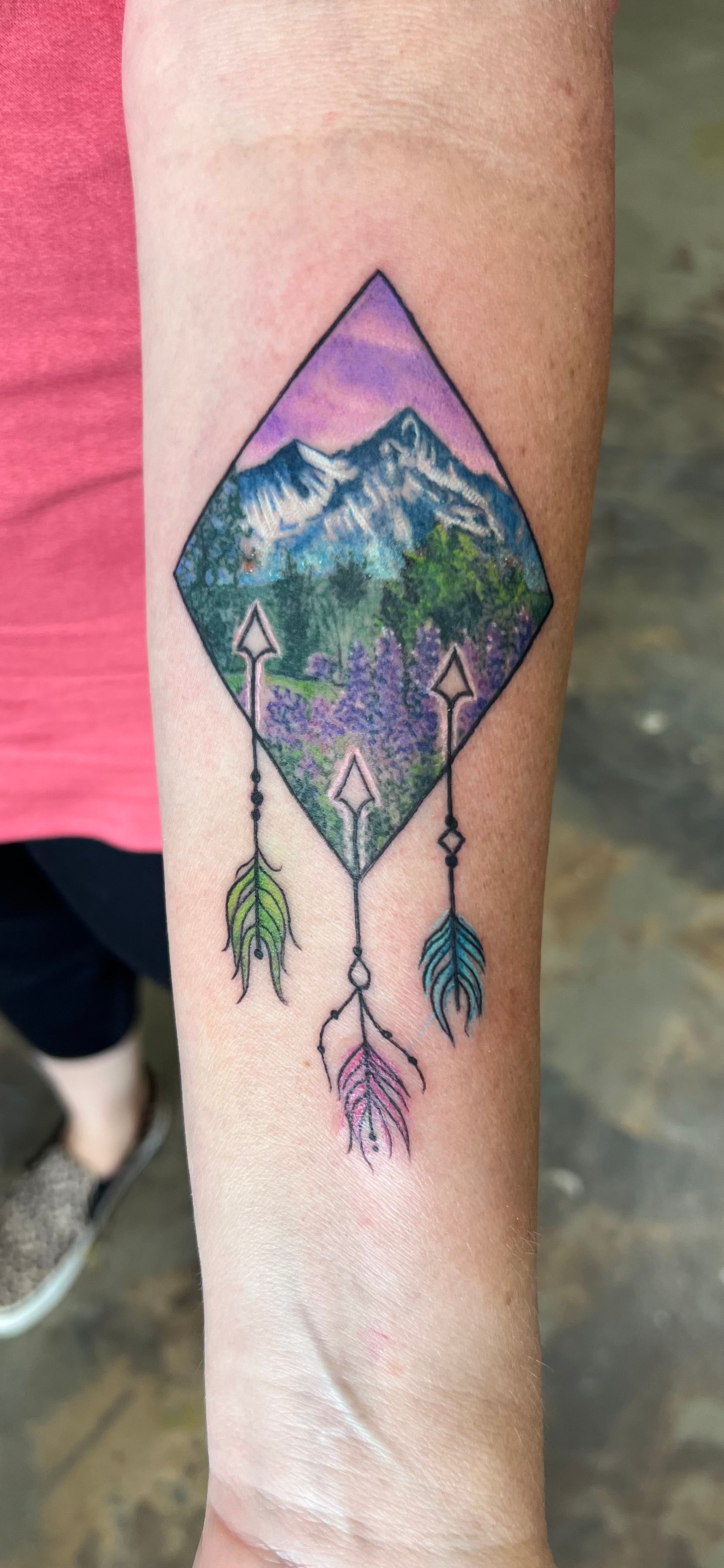 Tattoo of a diamond-shaped mountain scene with arrows and feathers, on a person’s forearm.