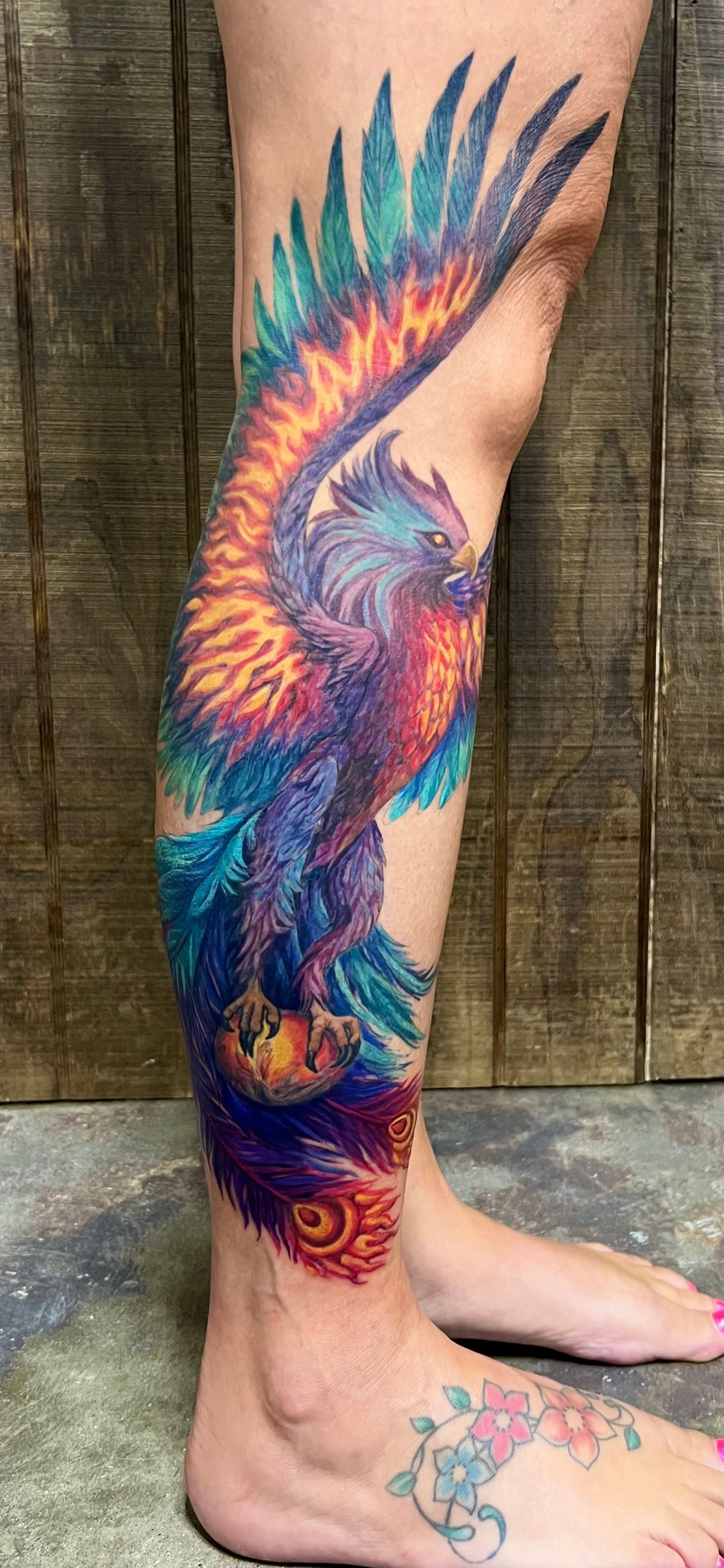 A colorful phoenix tattoo on a leg, with a floral tattoo on the foot.