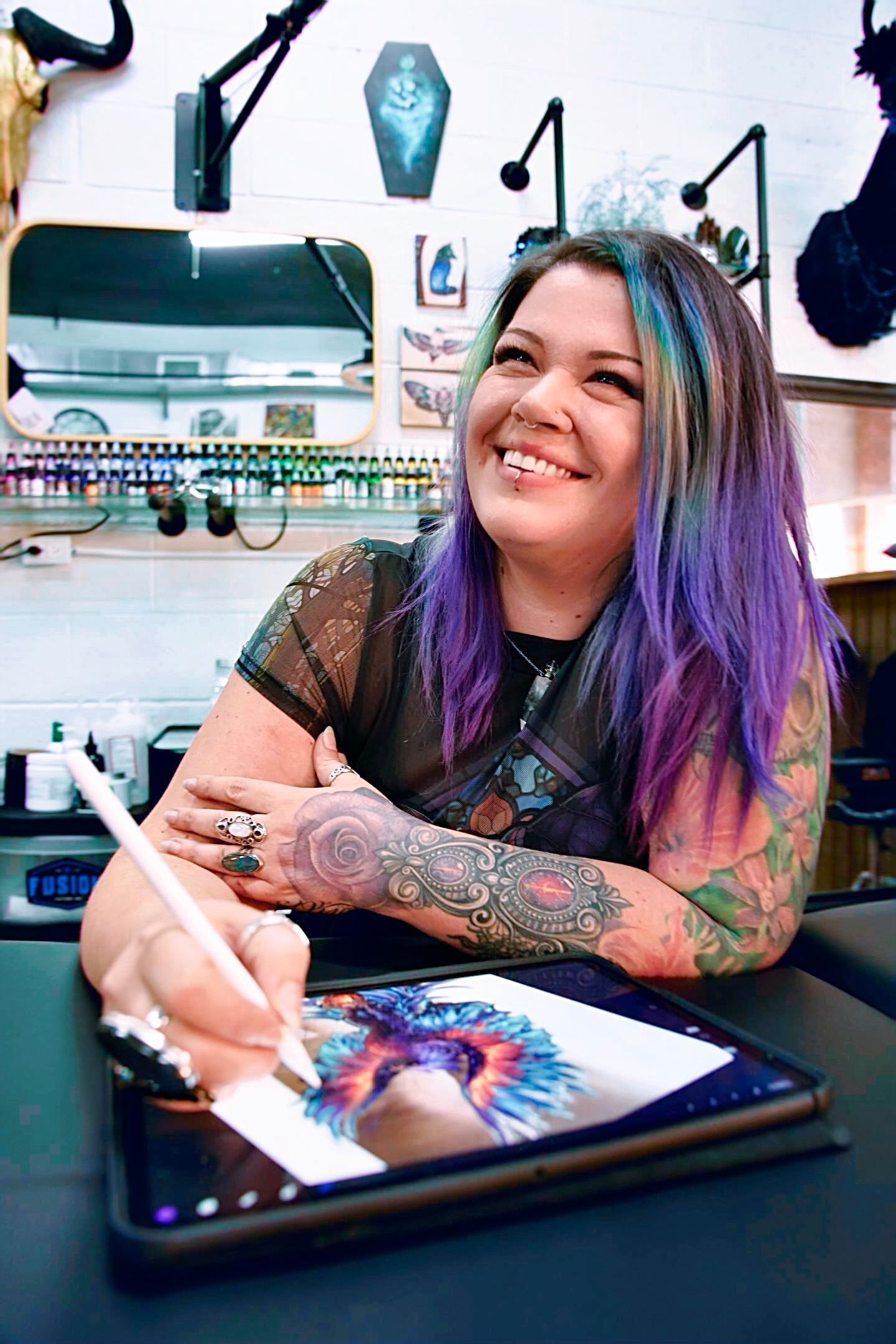 Woman with colorful hair, tattoos, smiles while drawing on a tablet in a tattoo studio.