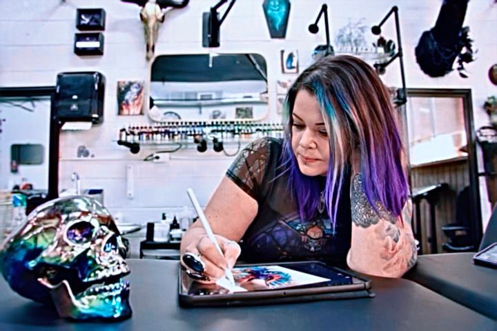 Woman with purple hair designs on a tablet at a tattoo shop, skull on table.