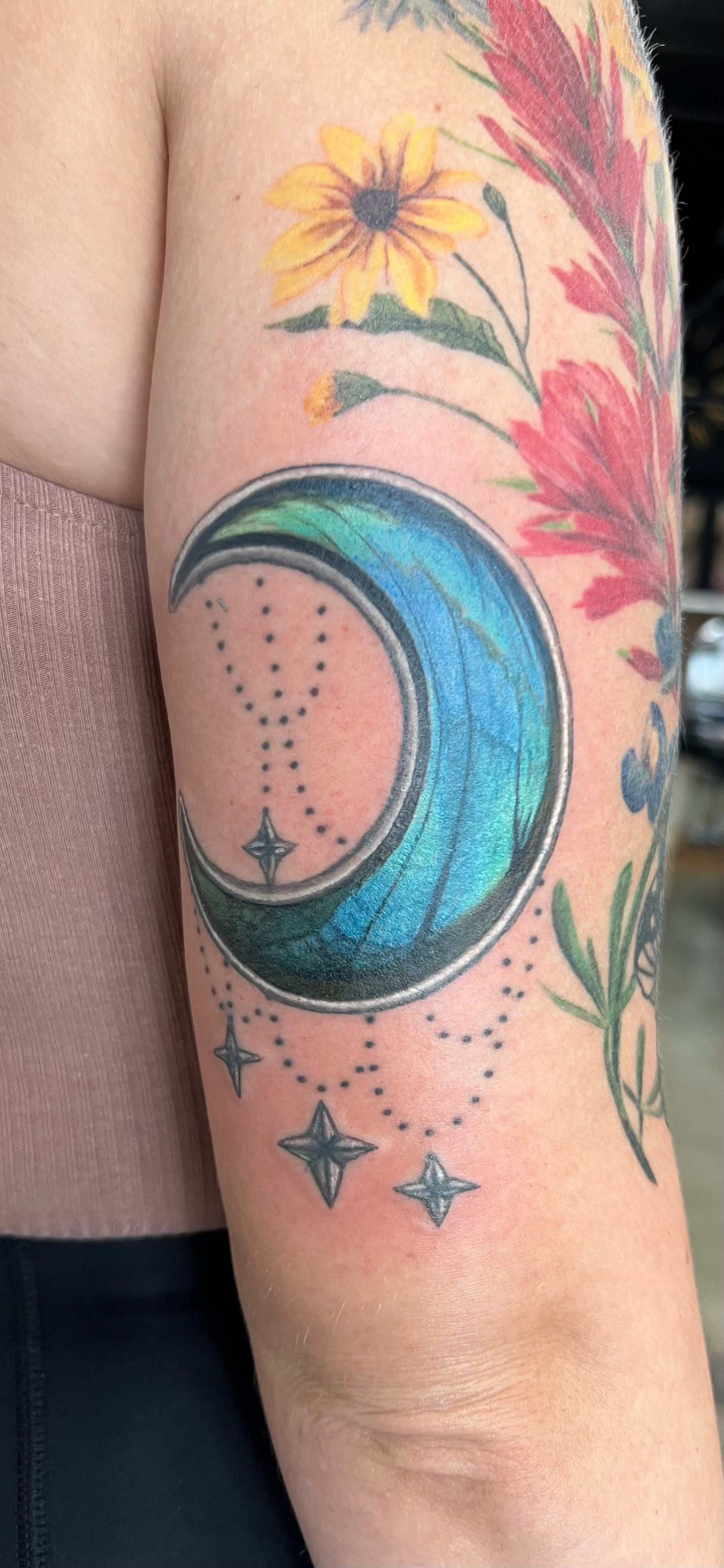 Tattoo of a crescent moon with a blue-green center and stars on an arm, with floral elements.