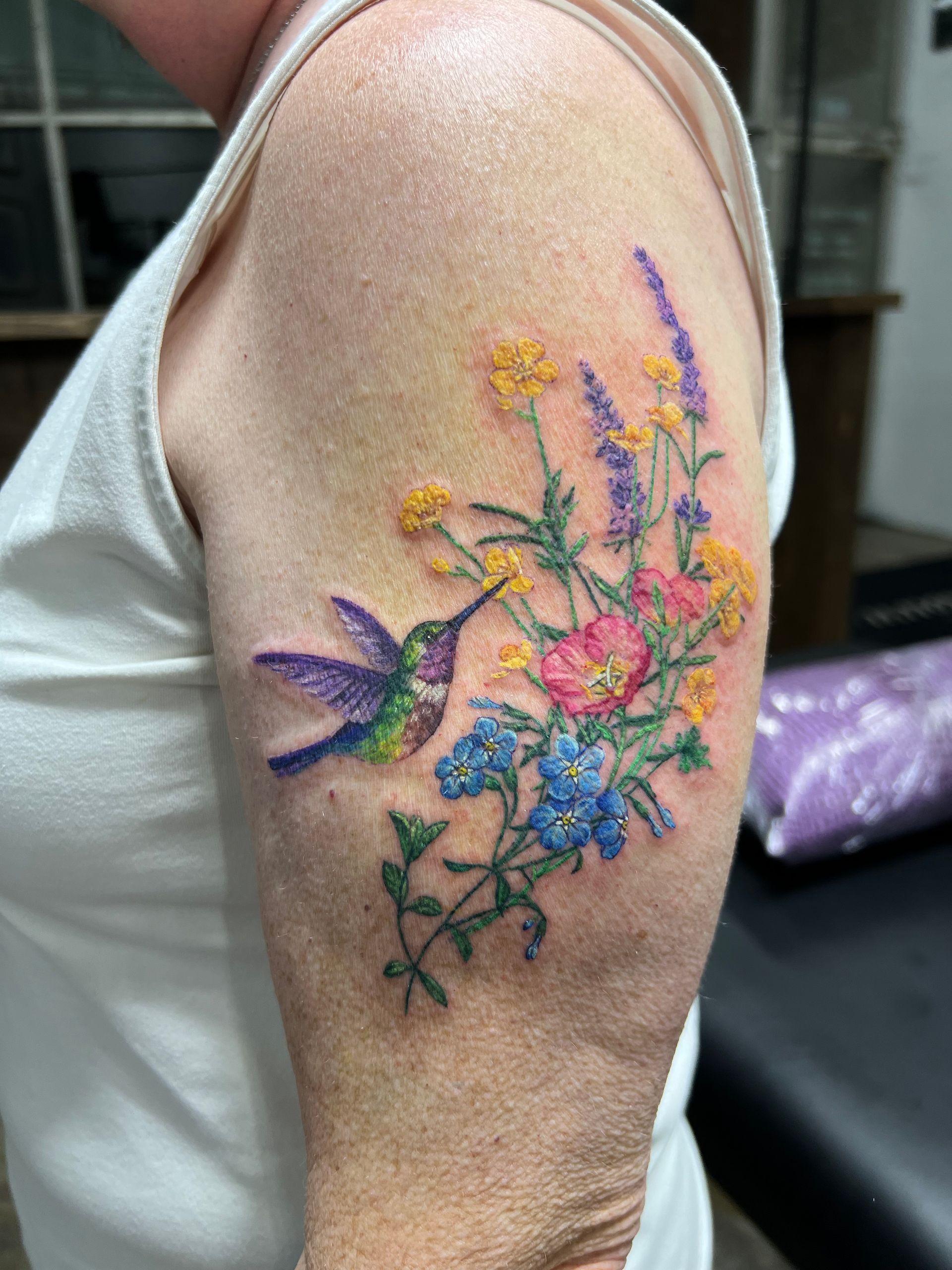 Tattoo of a hummingbird and flowers on a person's arm. Colorful bird drinking nectar from blooms.