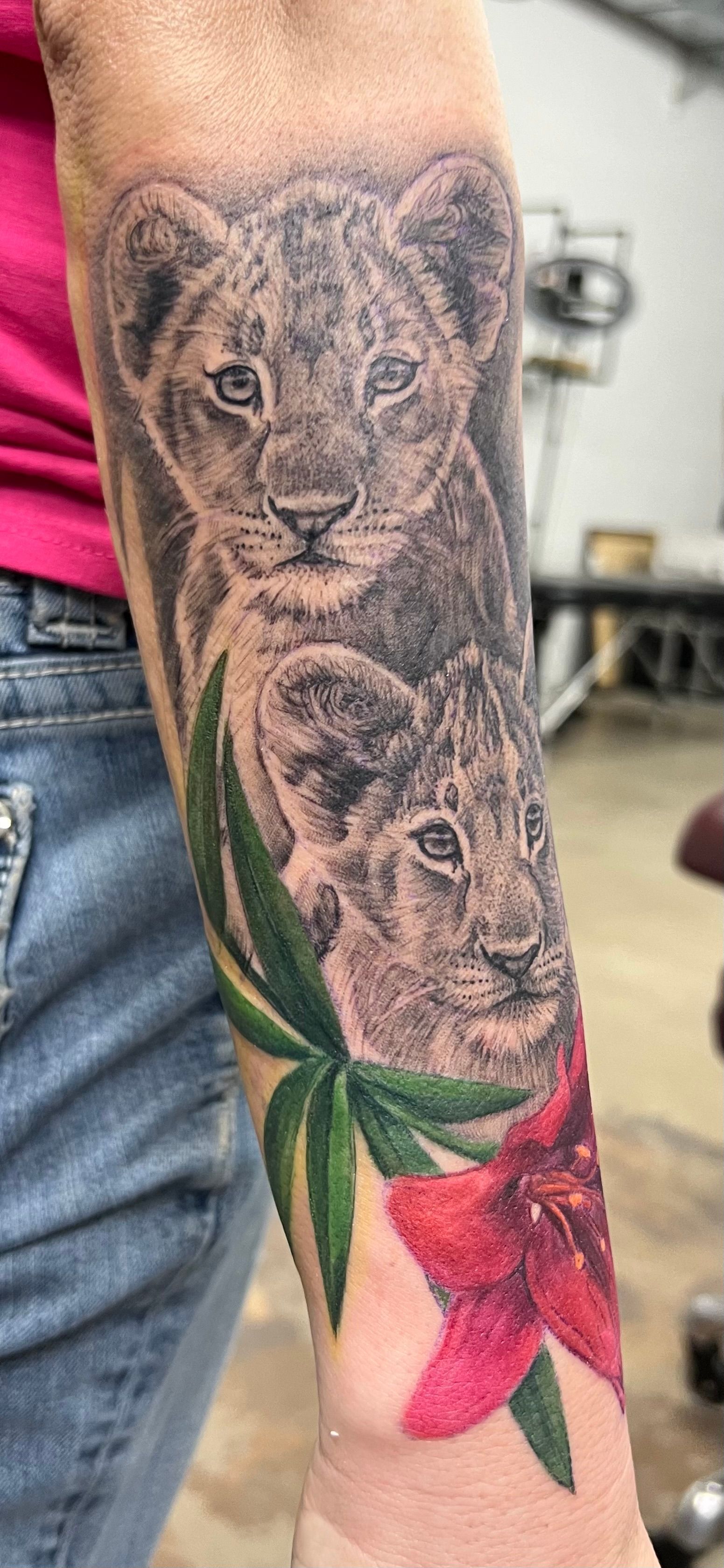 Tattoo of two lion cubs with greenery and a red flower on an arm.