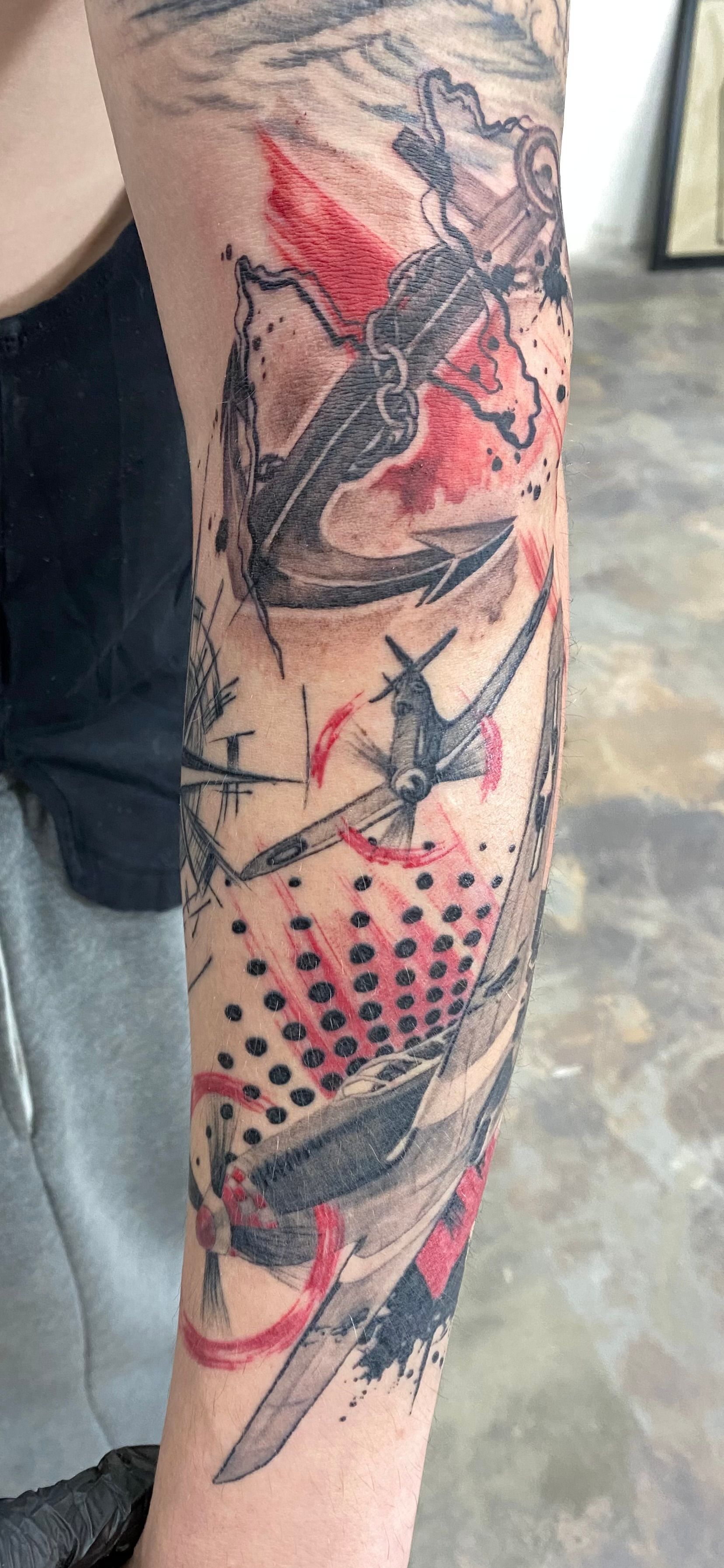 Tattoo of an anchor, plane, and stylized red and black shapes on an arm.