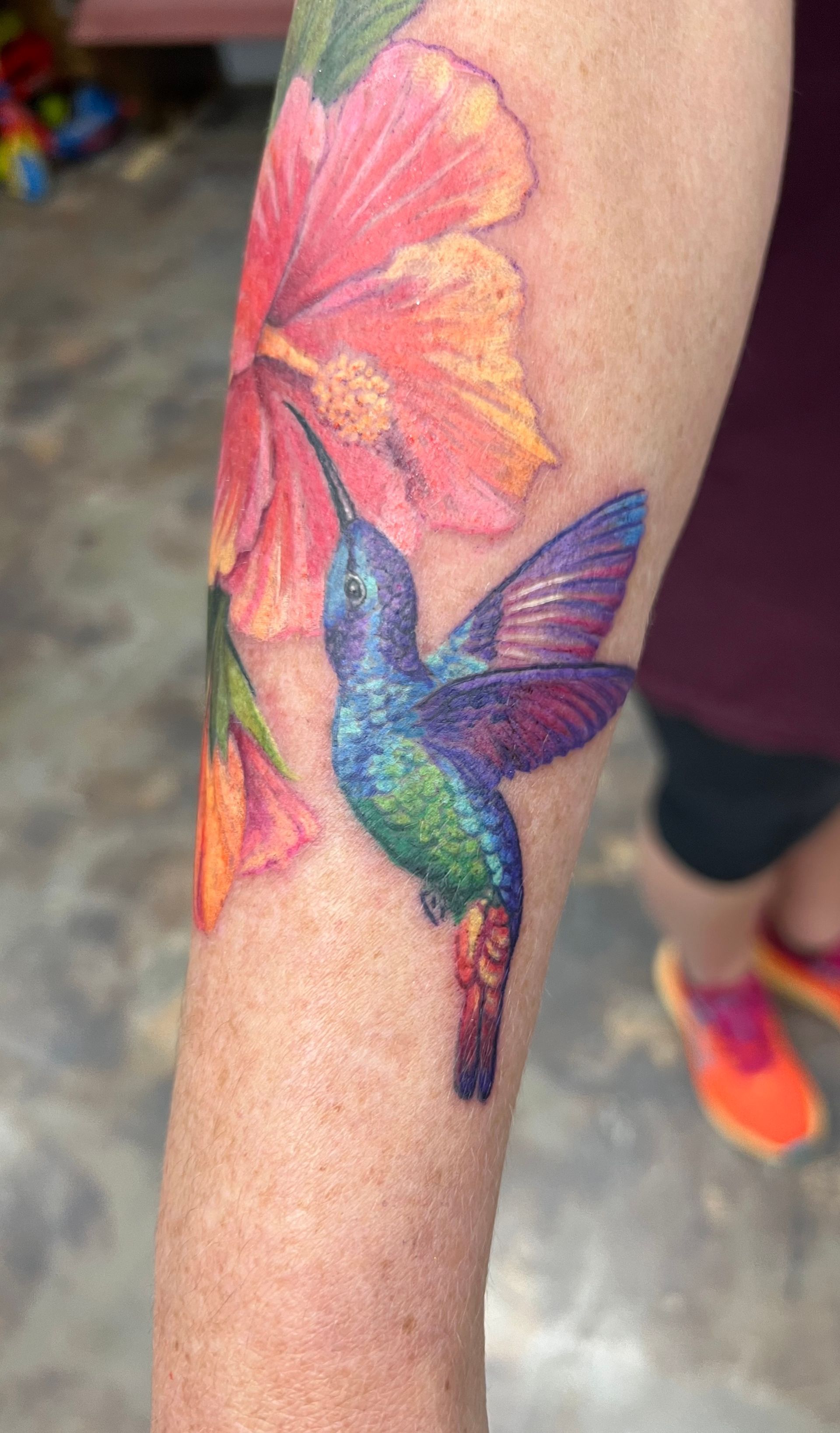 Hummingbird tattoo, iridescent blues/greens, feeding from orange/pink hibiscus. Arm.